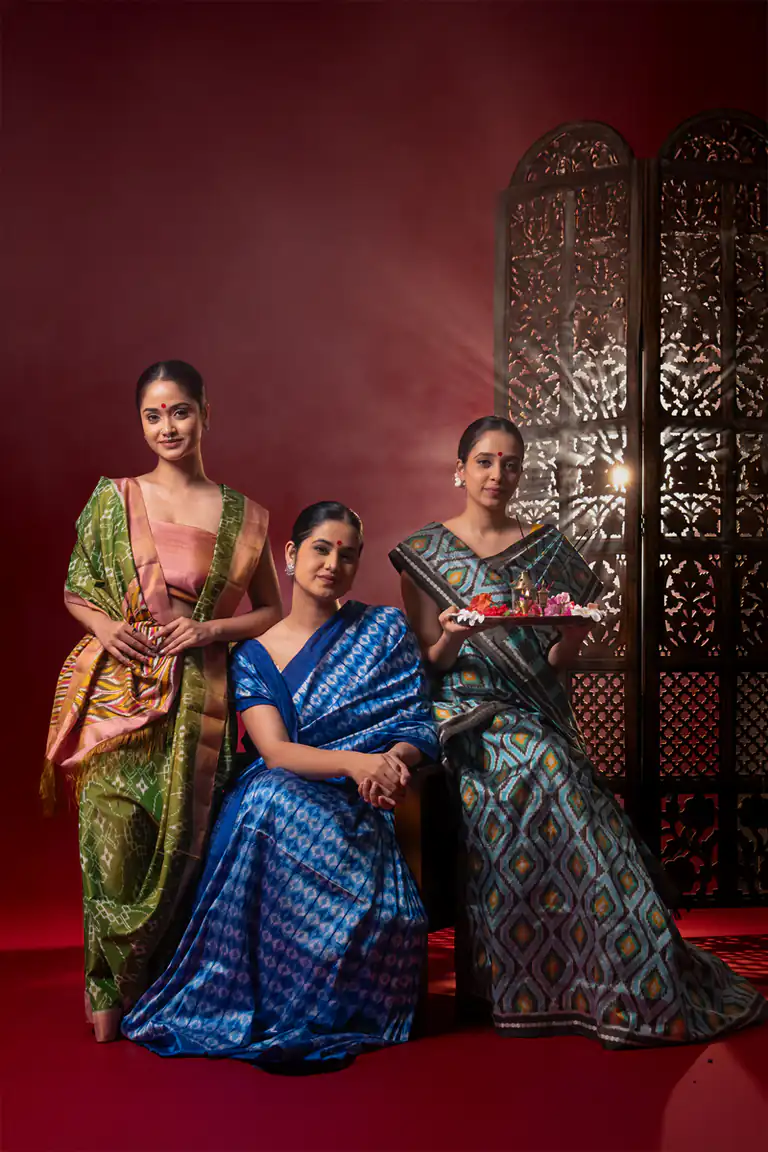 Three models wearing jewel-toned ikat saris posed in a deep red studio set